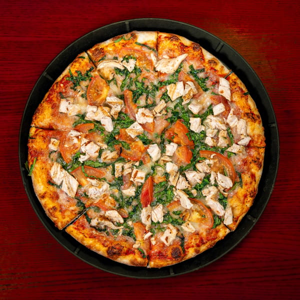 Overhead view of a chicken, tomato, and spinach pizza on a black pan, on a red wooden table.