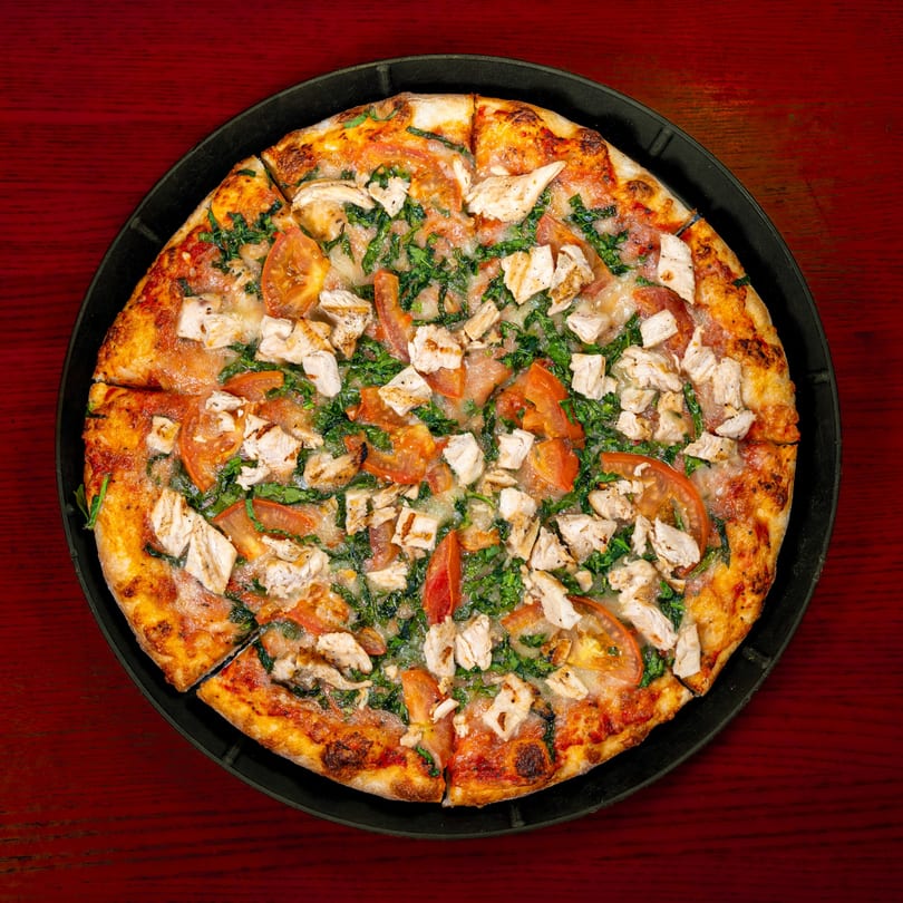 Overhead view of a chicken, tomato, and spinach pizza on a black pan, on a red wooden table.