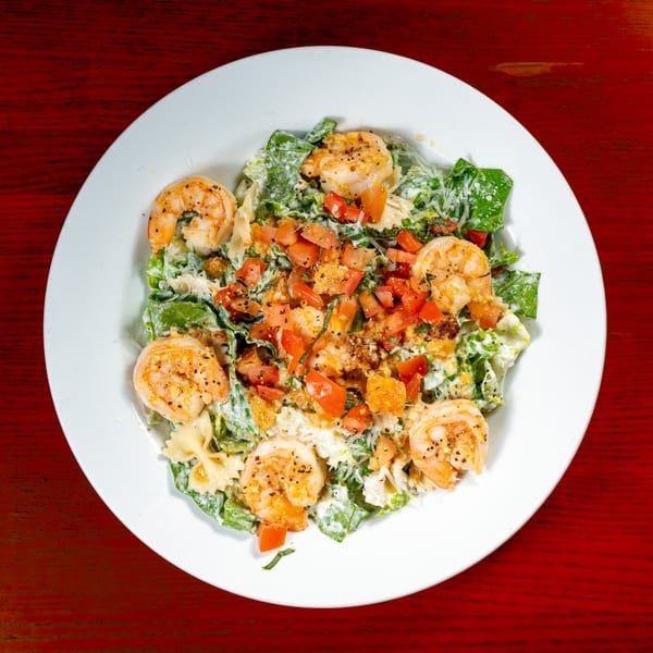 Overhead view of a shrimp salad with bowtie pasta, tomatoes, and greens on a white plate.