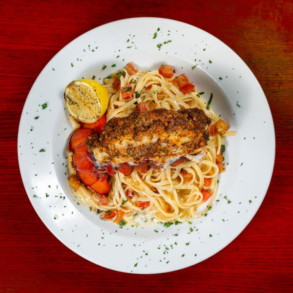 Lobster tail with linguine pasta, diced tomatoes, and a grilled lemon on a white plate.