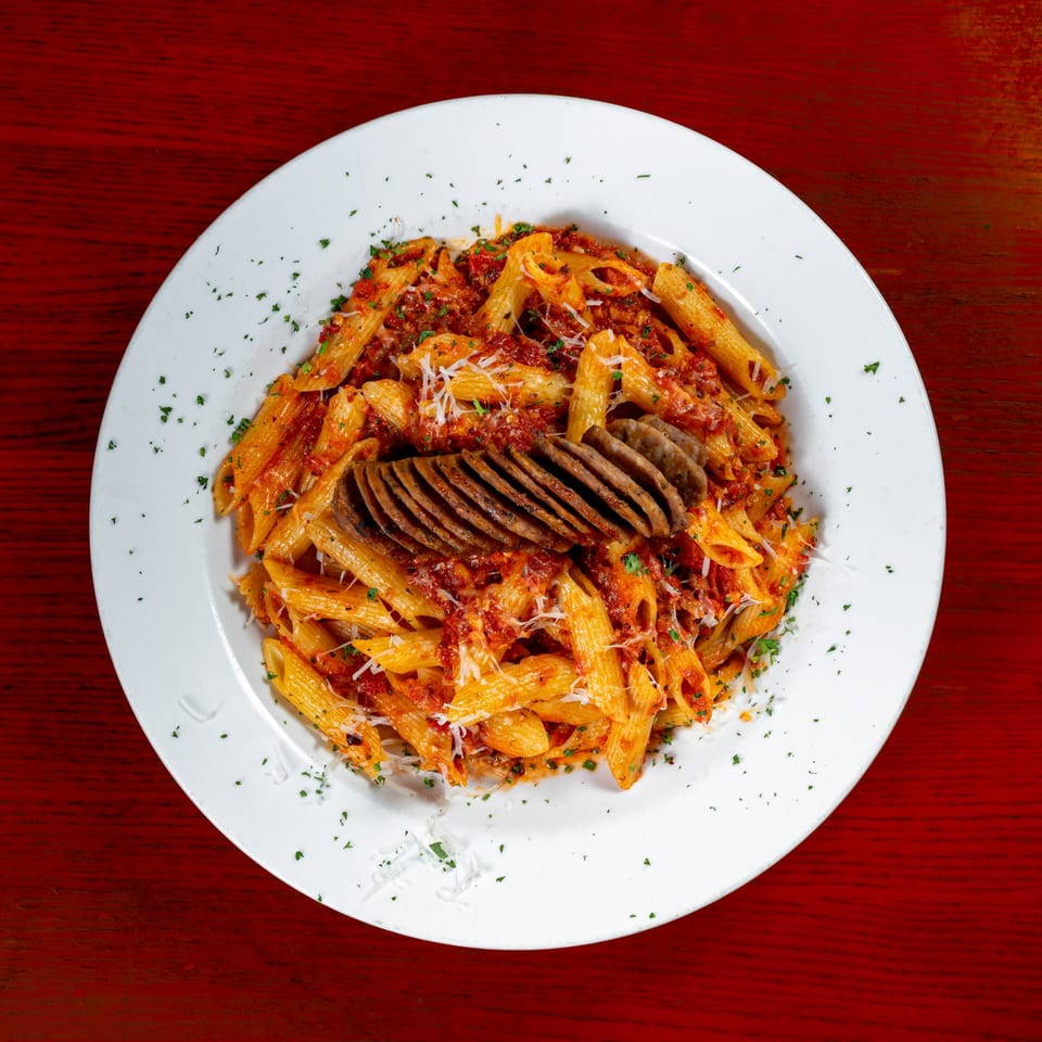 Penne pasta with red sauce, grated cheese, parsley, and sliced sausage on a white plate.