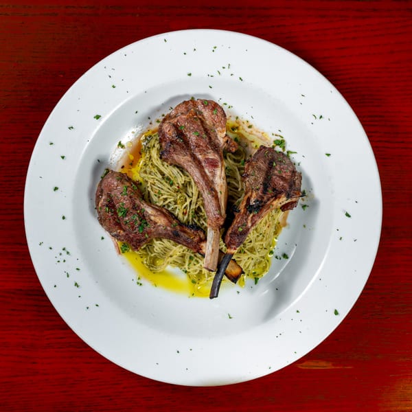 Three grilled lamb chops with pesto spaghetti on a white plate, garnished with herbs.
