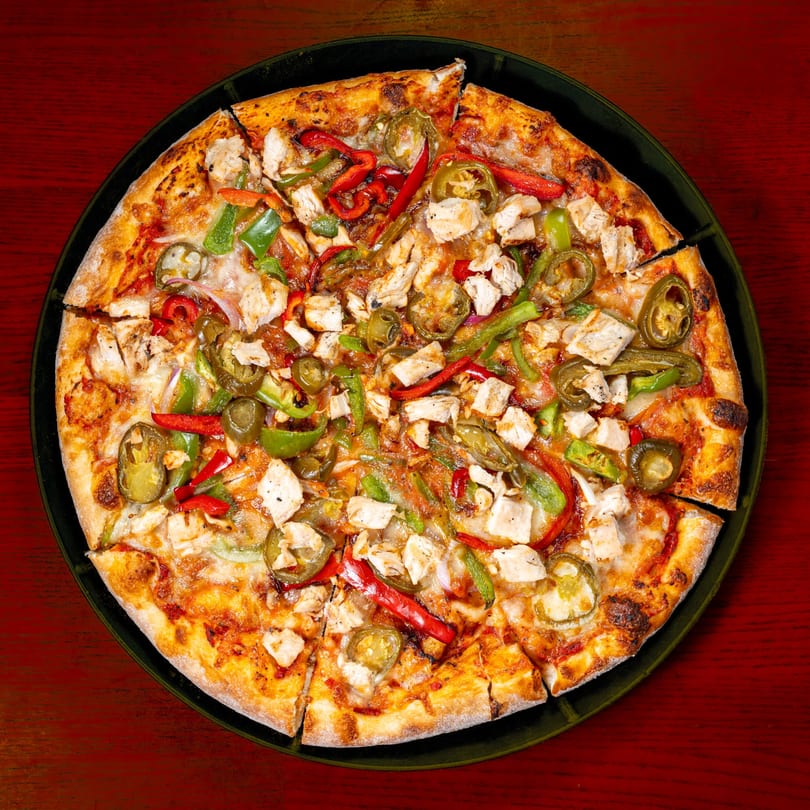 A whole chicken pizza with red and green peppers, and jalapeños, on a dark plate.