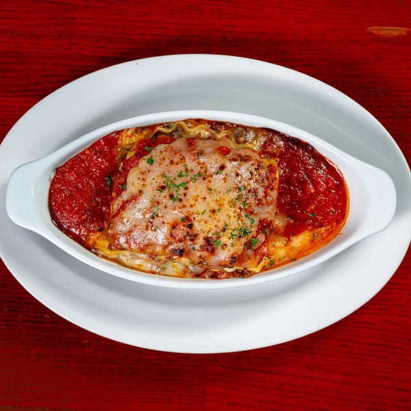 Baked lasagna with melted cheese and red sauce in a white oval dish on a red table.